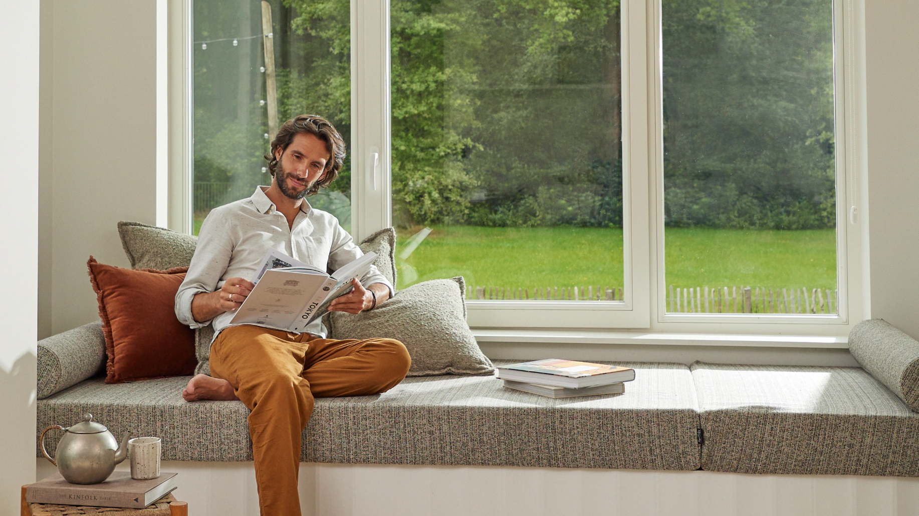 Άντρας που διαβάζει δίπλα σε ένα μεγάλο παράθυρο σε ένα σπίτι με ευχάριστη ατμόσφαιρα, δείχνοντας την άνεση και την αποδοτικότητα ενέργειας που παρέχουν οι αντλίες θερμότητας για να μειωθεί η επίδραση στο περιβάλλον σε λύσεις θερμότητας εσωτερικών χώρων