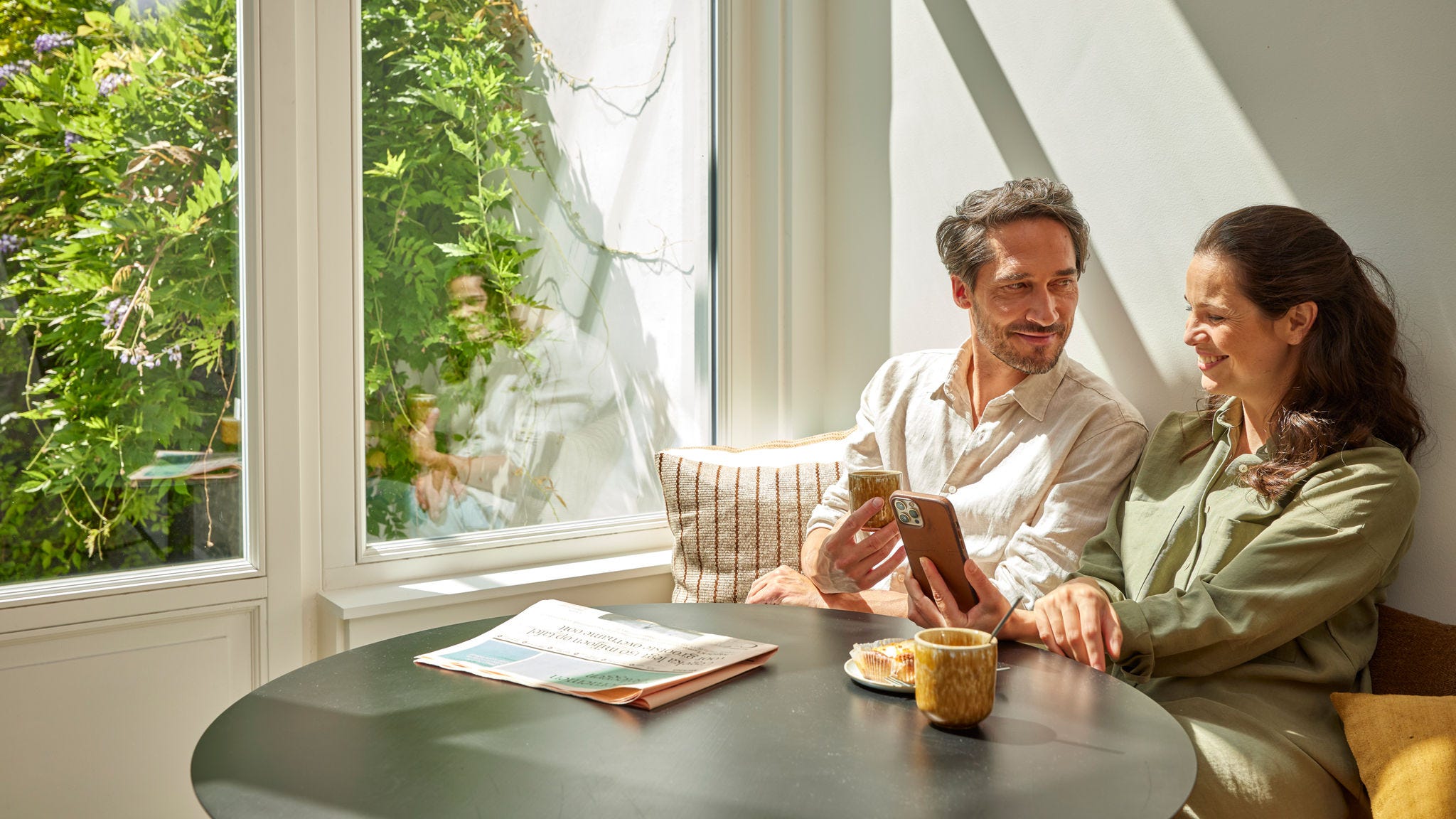 Ένα ζευγάρι απολαμβάνει τον καφέ του και βλέπει στην οθόνη του τηλεφώνου την εξοικονόμηση που πέτυχε από την ενέργεια, υποδηλώνοντας τις μειώσεις στα κόστη χάρη στη χρήση μιας αντλίας θερμότητας