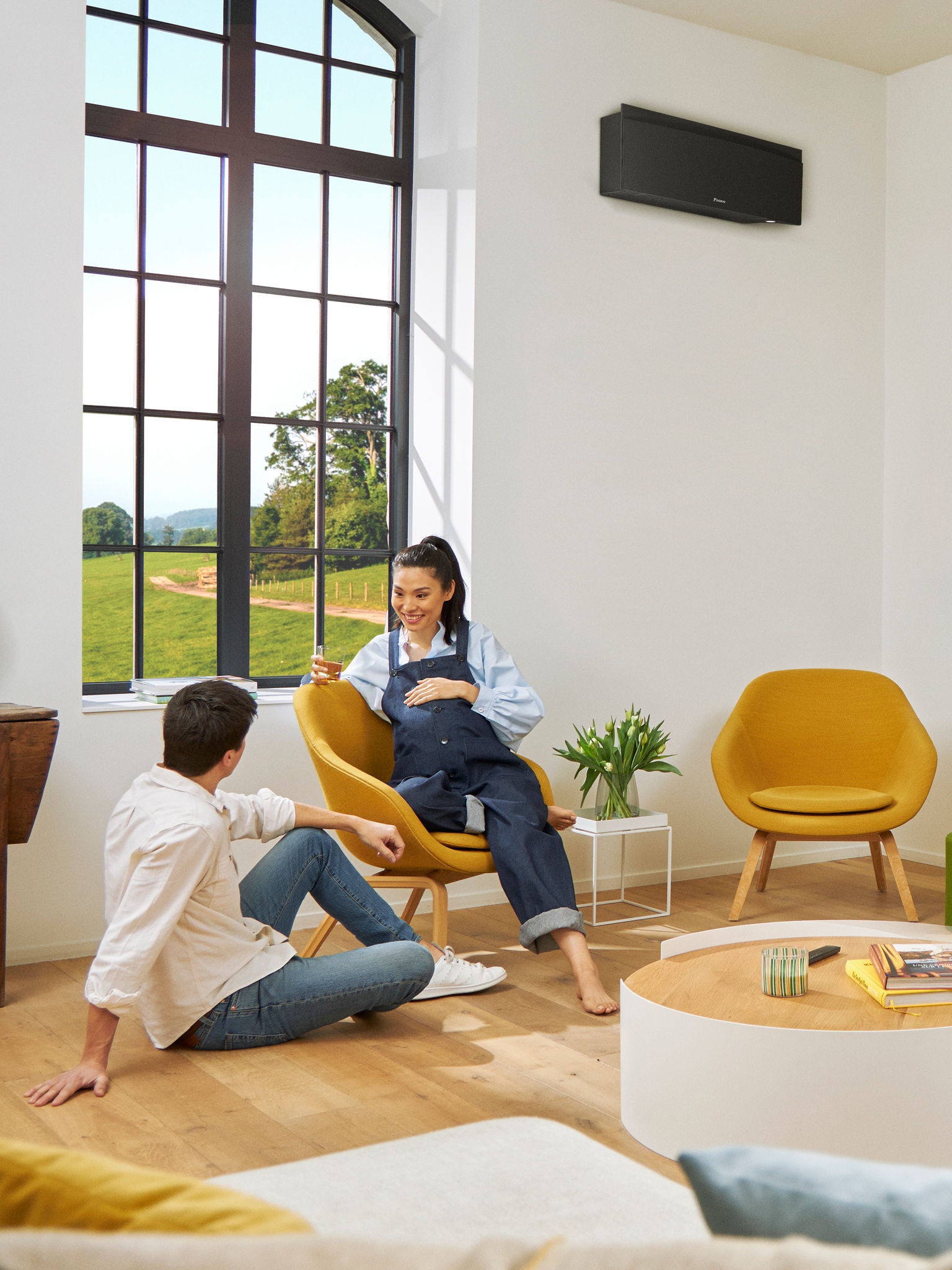 Ένας άντρας και μια γυναίκα κάθονται μπροστά από ένα μεγάλο παράθυρο σε ένα φωτεινό δωμάτιο και υπάρχει μια μαύρη επίτοιχη αντλία θερμότητας στον τοίχο πίσω τους