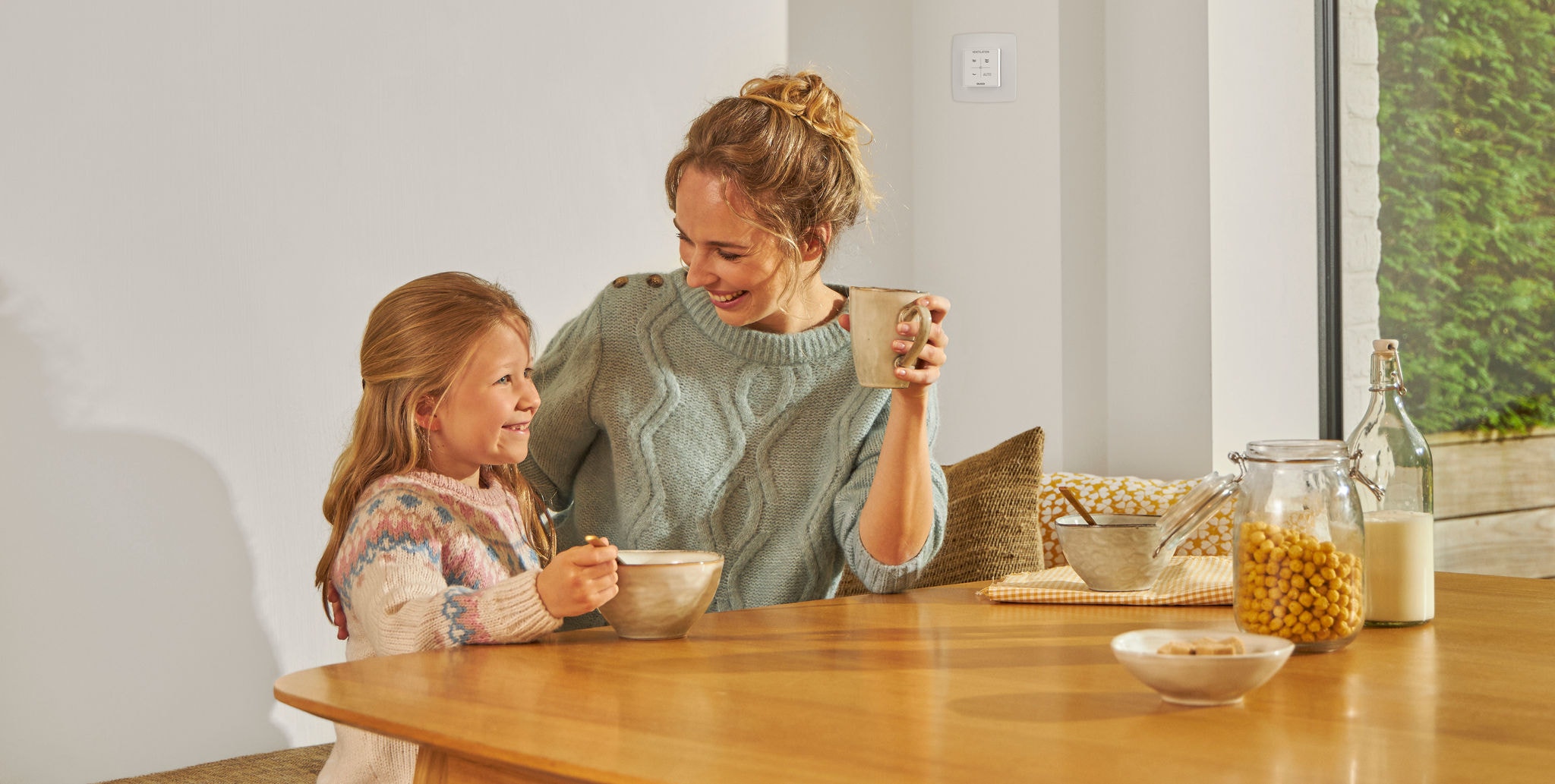 Νεαρό κορίτσι τρώει από ένα μπολ καθισμένη στο τραπέζι δίπλα σε μια γυναίκα που κρατάει μια κούπα, ενώ και οι δύο χαμογελούν, είναι χαρούμενες και χαλαρές, και απολαμβάνουν υγιεινό αέρα.