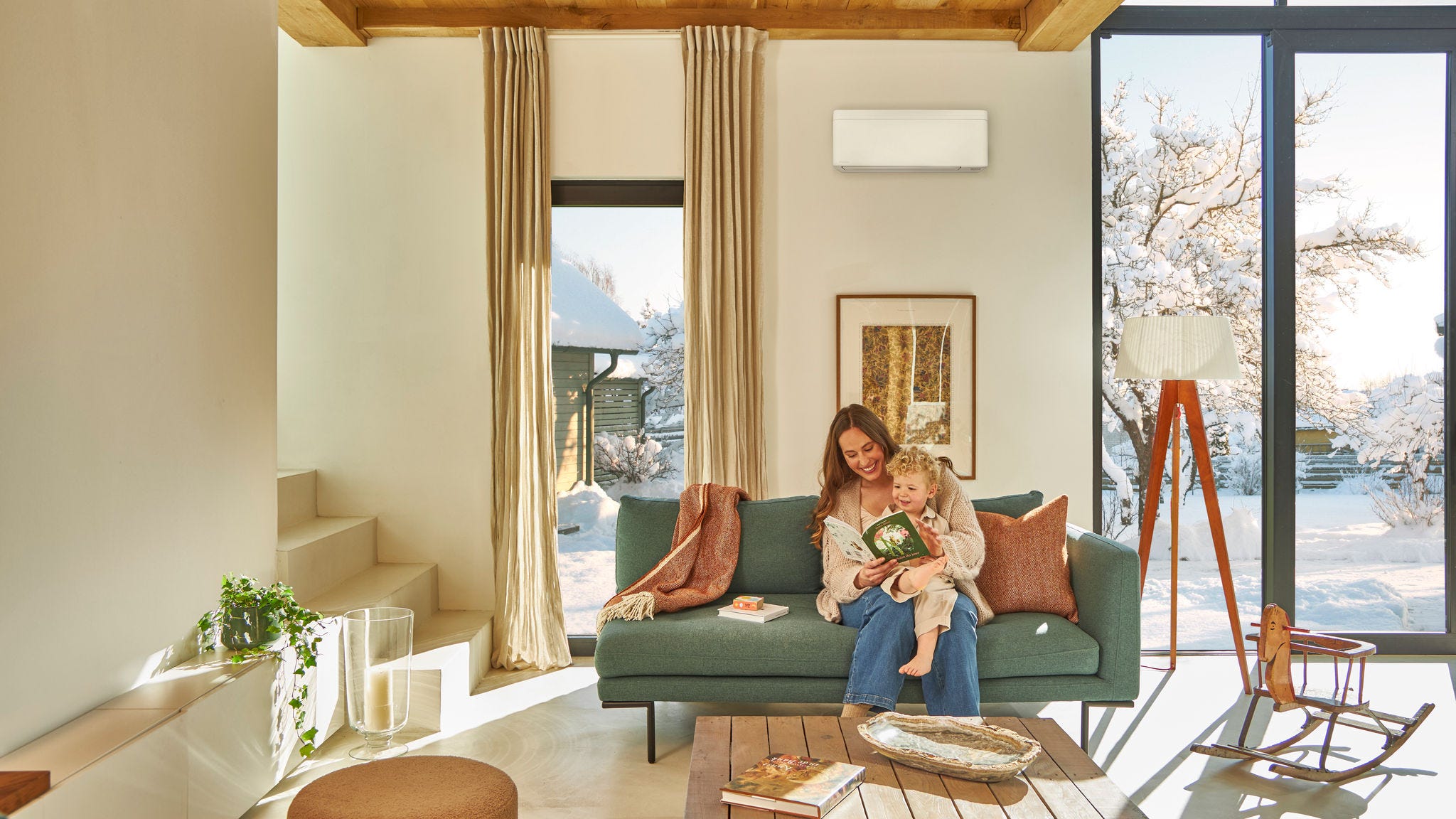 Μητέρα που κάθεται στον καναπέ κάτω από μια επιτοίχια αντλία θερμότητας αέρα-αέρα και διαβάζει ένα παιδικό βιβλίο με το παιδί της στα γόνατά της, απολαμβάνοντας τη ζεστασιά, ενώ έξω χιονίζει